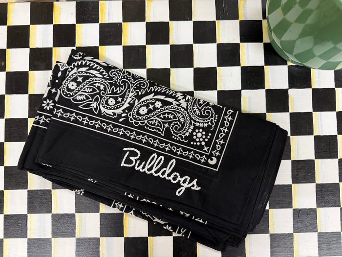 A black bandana-style cloth with white paisley print and the word 'Bullocks' in script, laid flat on a black-and-white checkered surface.