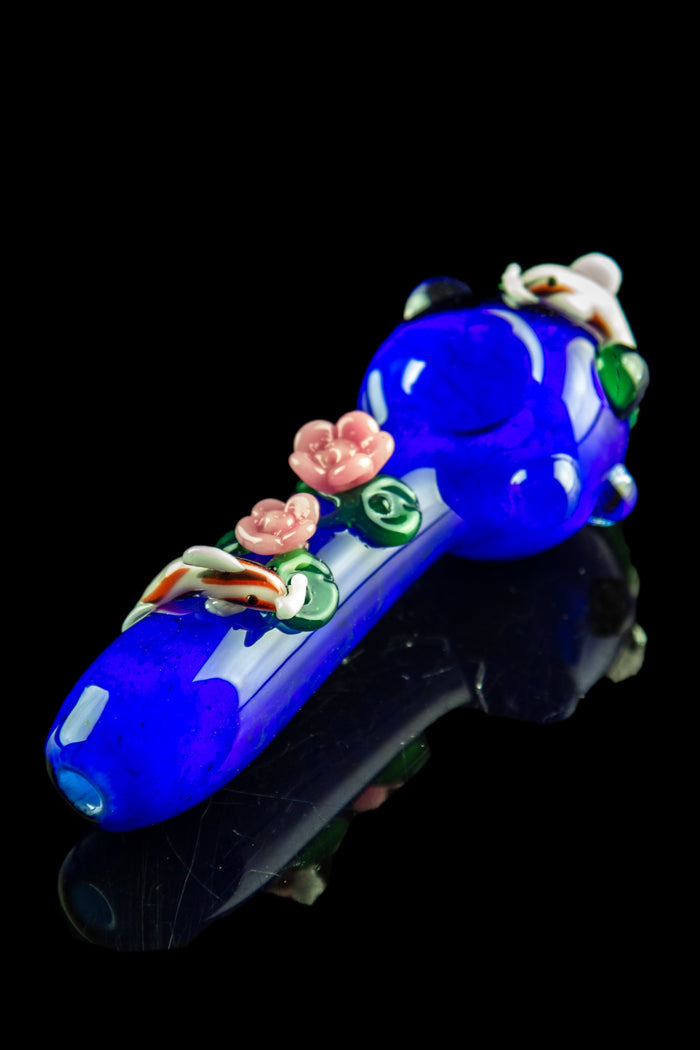 Close-up angled view of the Empire Glassworks "Koi Pond" Spoon Pipe showing its deep blue glass body with raised pink flowers and tiny koi accents.