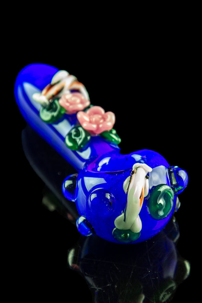 Close-up angled view of the Empire Glassworks "Koi Pond" Spoon Pipe in cobalt blue with raised green leaves and pink rose details on a black reflective surface