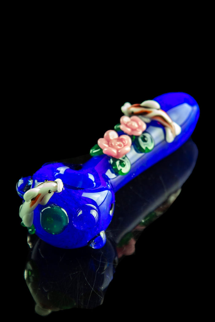 Angled close-up of the Empire Glassworks "Koi Pond" Spoon Pipe showing a cobalt blue glass body with raised pink floral and koi fish decorations on a black reflective surface