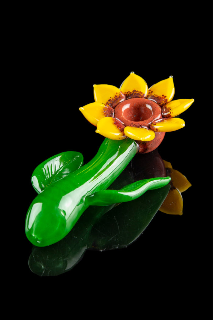 Hand-blown glass cannabis water pipe shaped like a blooming flower with green leaf accents, isolated on a black background.