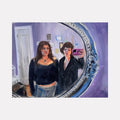 The artwork 'fit check before leaving Petra's' captures a mirror reflection in an ornate silver frame against a purple wall. Oil painting features soft brushstrokes depicting figures in black attire, with one holding a camera. Rich purple and blue tones dominate, with delicate details in the decorative mirror frame. Contemporary realist style. By Jillian Shea.
