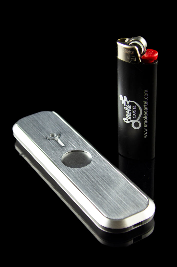Silver grinder and black lighter with “Smoke Cartel” logo on a reflective surface.