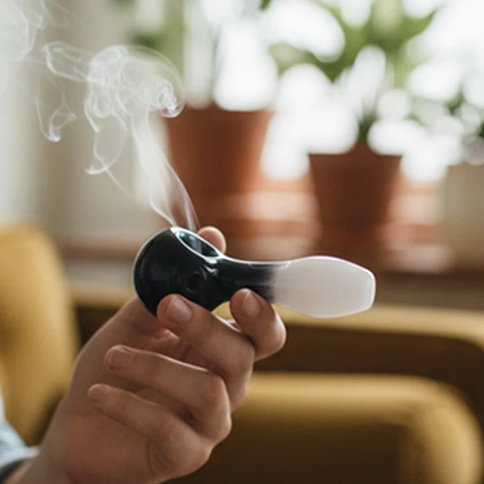 Close-up of a hand holding a smoking GRAV® Frit Spoon, shown in black-to-white glass with curling smoke and blurred potted plants in the background