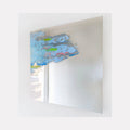 The artwork 'In Flux' by Sofia Echa features ethereal blue and green brushstrokes floating across polished stainless steel. Abstract forms reminiscent of clouds or water droplets create movement in the upper portion, contrasting with the reflective metallic surface. The 24x24 inch acrylic painting demonstrates a dynamic interplay between transparent colors and mirror-like material by Sofia Echa.