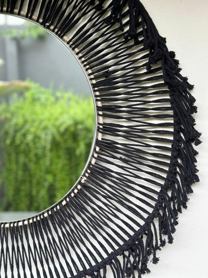 A round, black fringed hanging mirror with a visible pattern of crochet yarns.