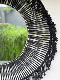 A round, black fringed hanging mirror with a visible pattern of crochet yarns.