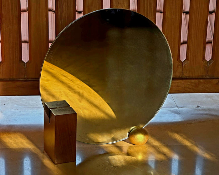A round, reclaimed teak wood mirror with a geometric design, reflecting a portion of a wooden structure and an orange object, placed on a flat surface.
