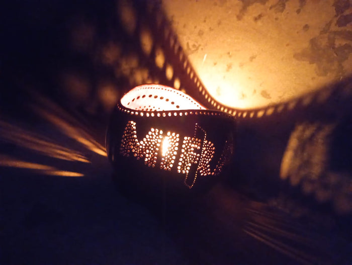 A personalized tea light holder with a ghee candle lit inside, placed on a surface with a shadow falling over it. The holder appears to be made of coconut shell and is customized with the word 'MIRAL'