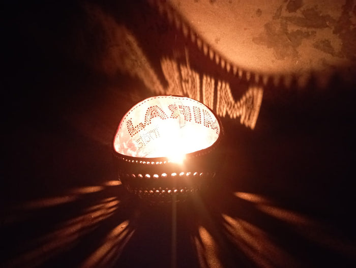 A personalized tea light holder with a ghee candle lit inside, placed on a surface with a shadow falling over it. The holder appears to be made of coconut shell and is customized with the word 'MIRAL'