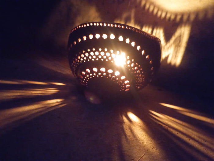 A personalized tea light holder with a ghee candle lit inside, placed on a surface with a shadow falling over it. The holder appears to be made of coconut shell and is customized with the word 'MIRAL'