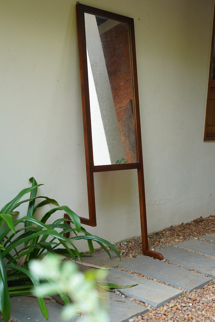 A handcrafted floor-standing mirror with a leaning section, made of Saint Gobain glass and Nilambur teak wood, positioned on a stone floor with a plant beside it.