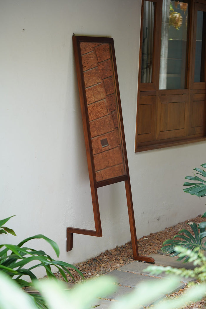 A handcrafted floor-standing mirror with a leaning section, made of Saint Gobain glass and Nilambur teak wood, positioned on a stone floor with a plant beside it.