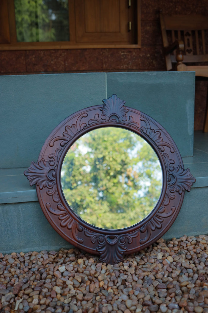 A circular teak wood mirror with floral carving patterns, displayed against a white wall with decorative items to the side.
