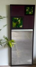An asymmetrical wall-mounted mirror with a black frame, featuring a live moss abstract art section on one side and a rectangular mirror on the other, set against a wall with a large teak wood door.