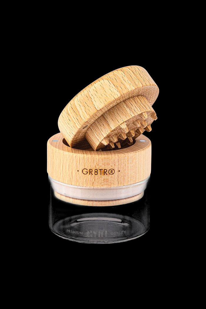 A close-up, angled view of the RYOT Beech Wood GR8TR Grinder with Glass Jar, showing the wooden lid and clear glass base.