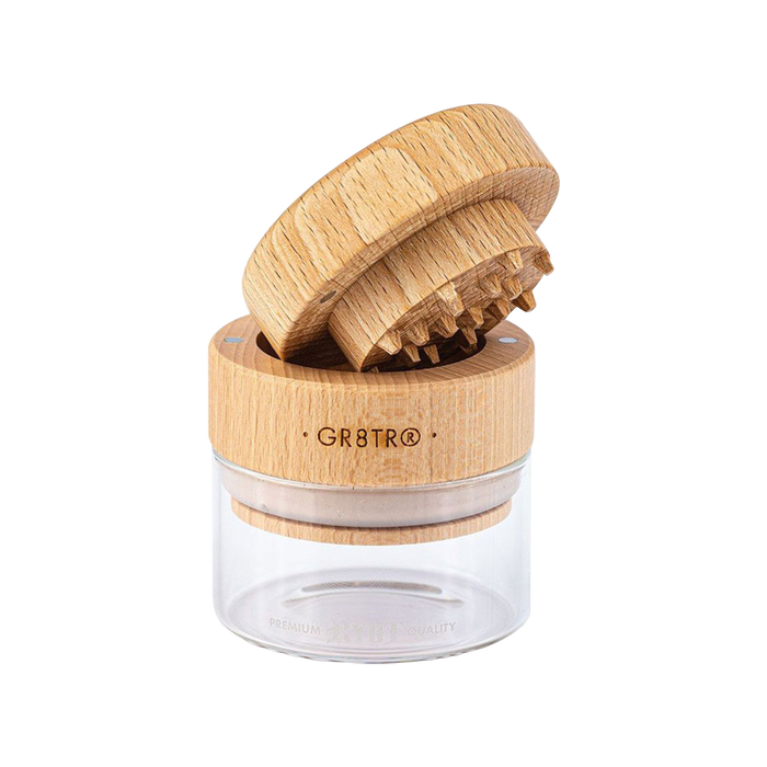 A close-up, top-down view of the RYOT Beech Wood GR8TR Grinder with Glass Jar, showing its textured wood lid and clear glass base.
