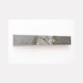 The artwork 'Sidewalk Crack' by Ryan Frank captures a minimalist urban landscape detail, featuring a diagonal fissure in concrete pavement with small green weeds emerging through. The color photograph, mounted on sapele wood, presents a stark contrast between gray concrete texture and resilient plant life, measuring 2.5 x 16 inches.