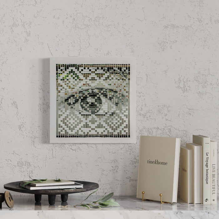 The artwork 'Warning' by Kiana Honarmand features a pixelated eye pattern created through hand-cut and woven giclee prints. The composition displays a striking mosaic-like effect in black, white, and subtle green tones, arranged in a geometric pattern within a square white frame. The 16x16 inch piece combines digital aesthetic with traditional weaving techniques by Kiana Honarmand.